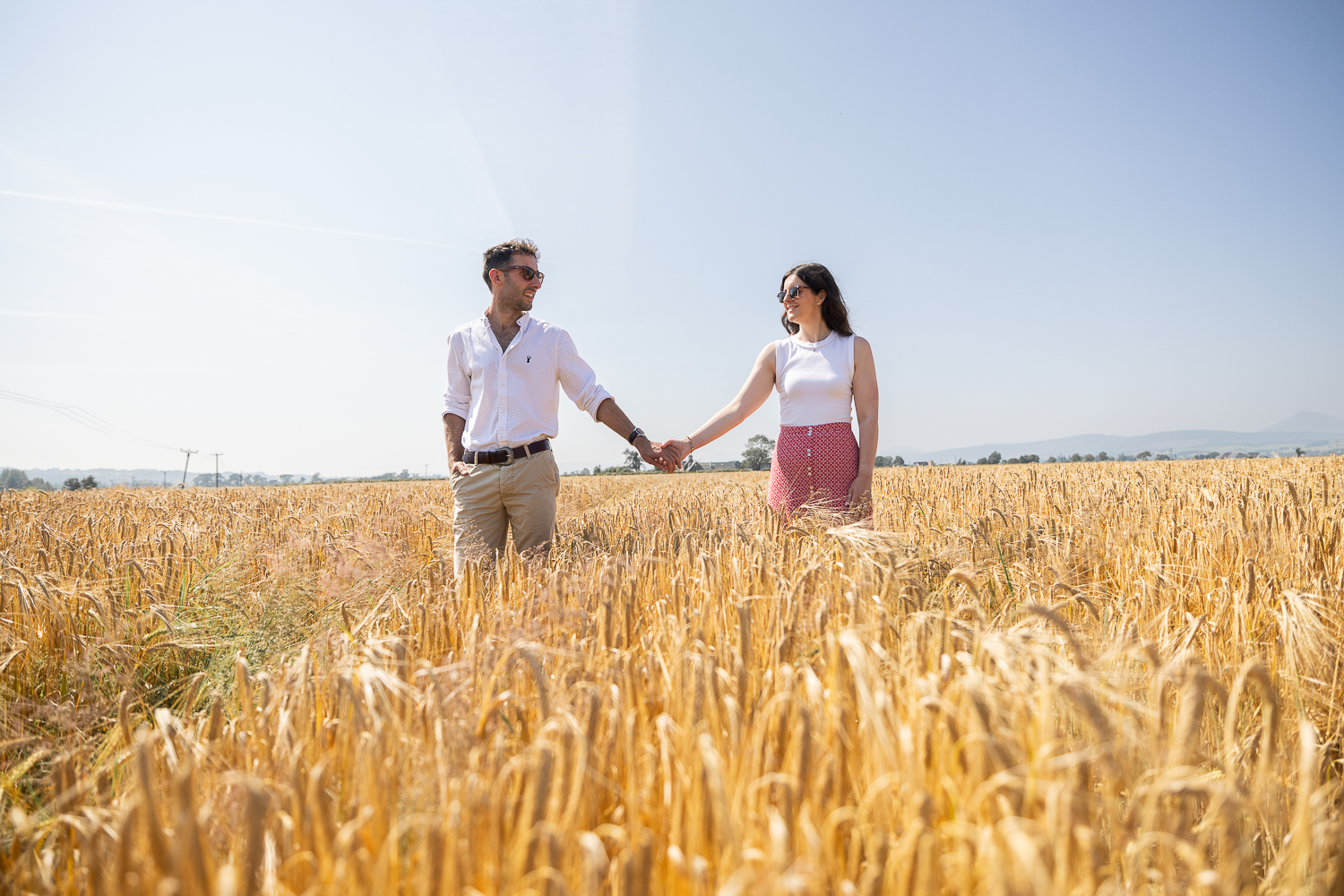 Emma and David holding hands in a golden wheat field on David's family farm in Stirlingshire.